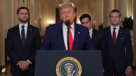 Trump, wearing a blue suit, is flanked by three officials as he delivers his address to the nation at a lectern